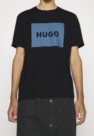 Person iført en sort T-shirt med korte ærmer med et blåt rektangulært logoblok, der viser "HUGO" med sorte store bogstaver.