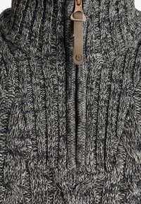 Gebreide trui in grijs en zwart met een hoge kraag, voorzien van een rits aan de voorkant en een leren trekker. Gedetailleerd met een kabelbreipatroon.