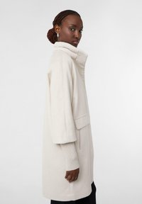 Cappotto beige chiaro con colletto alto, maniche a tre quarti e polsini a coste; presenta una tasca laterale e un tessuto morbido e testurizzato.