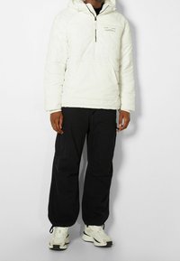 Vit puffjacka med dragkedja i kragen, svarta cargobyxor, och beige sneakers. Funktioner inkluderar sömdetaljer och en logotyp på bröstet.