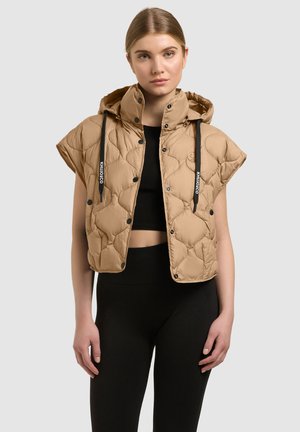 Mladá žena oblečená v béžovej bezrukávovej prešívanej kapucňovej veste s čiernymi sťahovacími šnúrkami cez čierny krátky top a legíny.