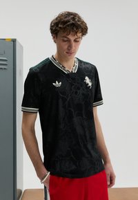 T-shirt de sport noir avec motif floral, col et poignets rayés verts et blancs. Arborant le logo Adidas sur la poitrine, assorti à un short rouge.