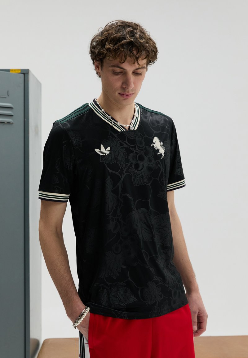 T-shirt de sport noir avec motif floral, col et poignets rayés verts et blancs. Arborant le logo Adidas sur la poitrine, assorti à un short rouge.