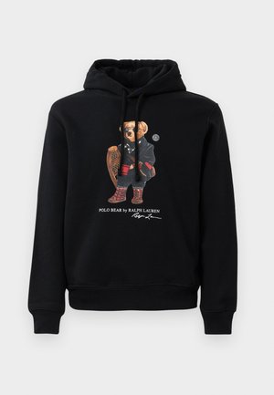 Hoodie noire avec un ours portant des lunettes de soleil, un manteau, des bottes et tenant une raquette en bois, avec le texte "Polo Bear by Ralph Lauren" en dessous.