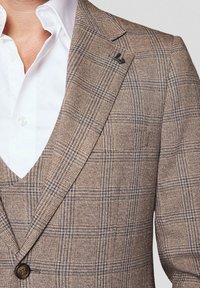 Brązowa marynarka w kratę z niebieskimi i beżowymi wzorami, z teksturowanej tkaniny, z klapami i czarnym detalem logo na klapie. Pod nią widać białą koszulę.