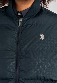 Ciemnozielona pikowana kurtka typu bomber z prążkowanym kołnierzem, ozdobiona subtelnym wytłaczanym wzorem oraz srebrnym logo na piersi. Zamykana na suwak.