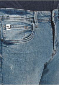 Primer plano de unos vaqueros de denim azul claro con una pequeña etiqueta blanca que dice "INDICODE" cerca del bolsillo frontal y una camisa oscura metida por dentro.