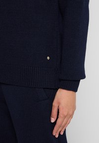 Navyfarbener Strickpullover mit gerippten Bündchen und Saum, versehen mit einem kleinen goldenen Logodetail an der Seite. Weicher Stoff und lockere Passform.