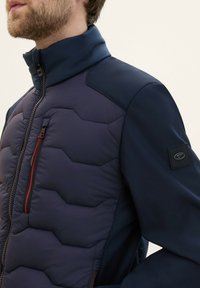 Mörkblå jacka med vadderad front som har ett hexagonalt mönster, hög krage och kontrasterande texturerade ärmar. Röd dragkedjedetalj är noterad.
