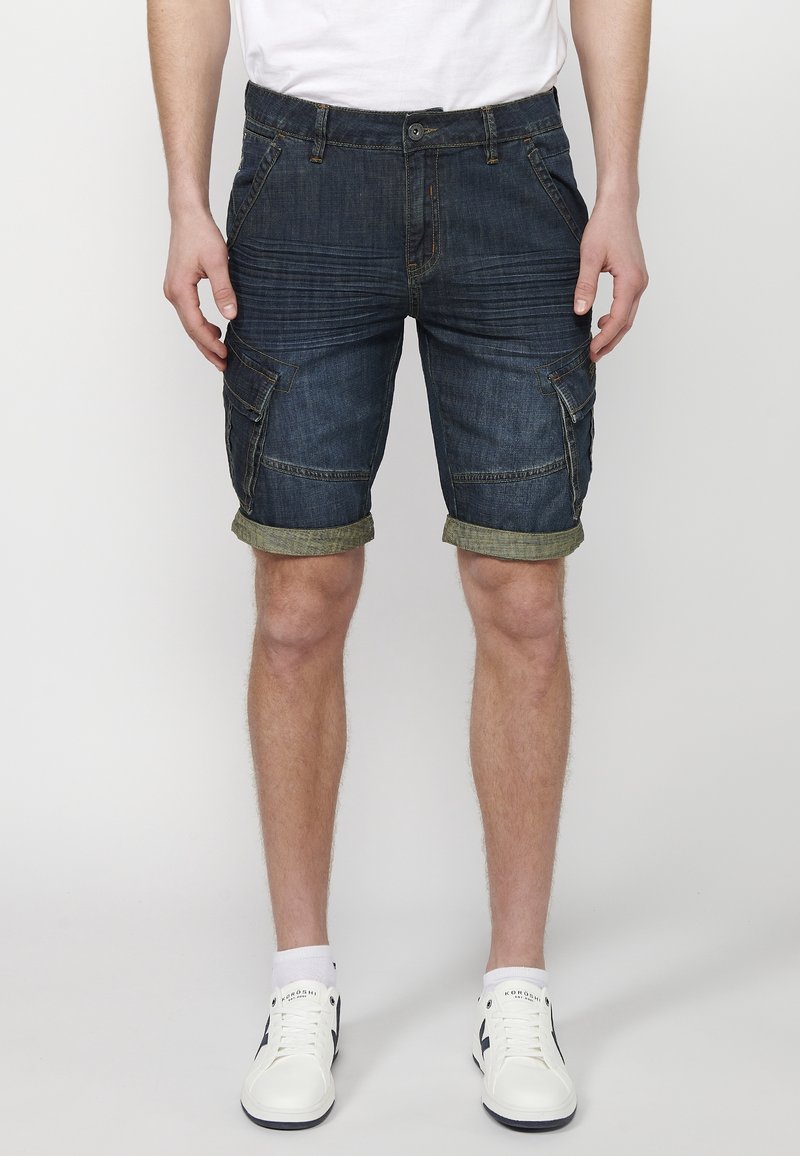 Homme portant un short cargo en denim bleu foncé retroussé, des baskets blanches et un t-shirt blanc, debout devant un fond blanc.