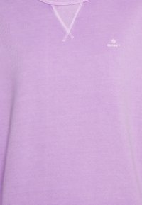 Sudadera de color lila claro con cuello redondo, que presenta un pequeño logo blanco. Textura suave y un ajuste relajado con detalles de costuras reforzadas.