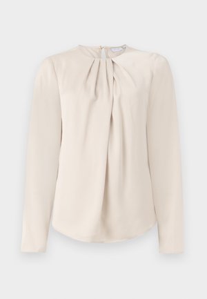 Blouse beige à manches longues avec un col rond, présentant des plis sur le devant et un ourlet arrondi. Tissu lisse avec une fermeture éclair invisible au dos.