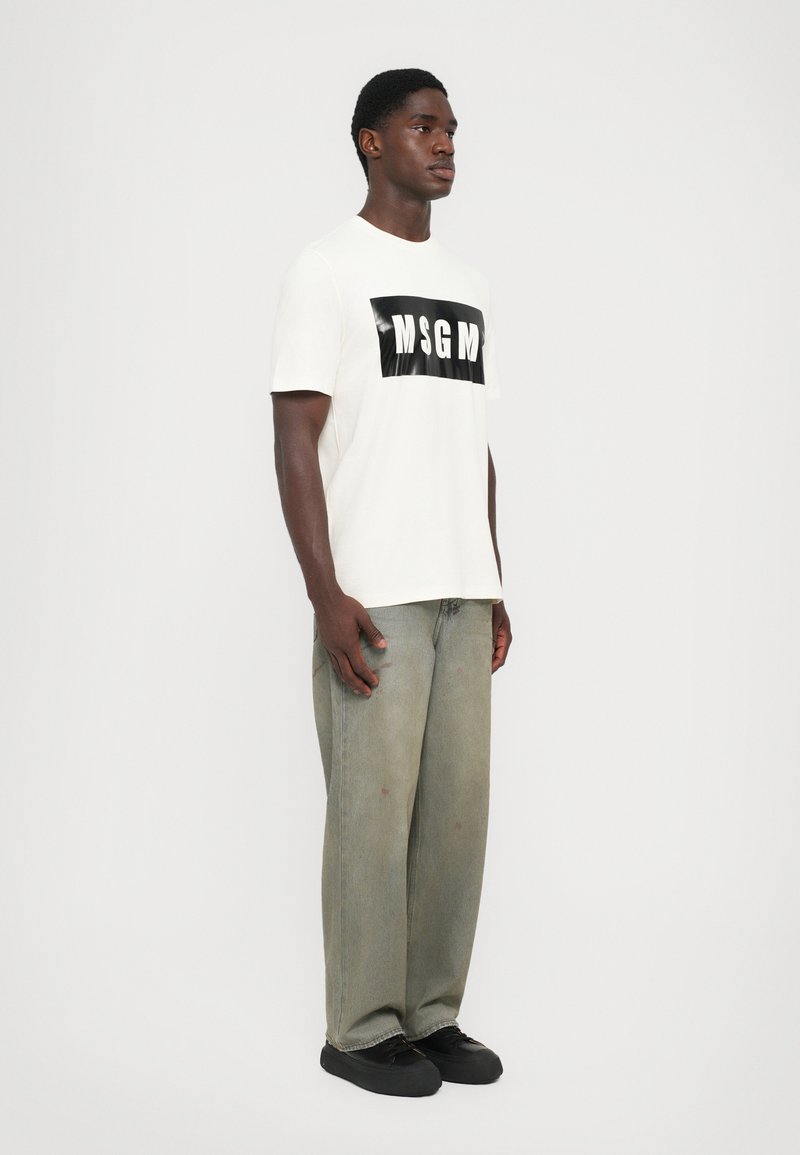 White cotton T-shirt with a black glossy MSGM logo centered on the chest, paired with oversized, faded green denim jeans and black shoes.