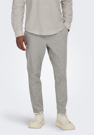 Mann trägt hellgraue, gerippte Hose, beiges Langarmshirt und cremeweiße klobige Sneakers, steht mit den Händen in den Taschen.