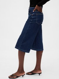 Osoba nosi tamnoplavih traperica culottes i crnih sandala s niskim potpeticama, s jednom rukom oslonjenom na kuk.