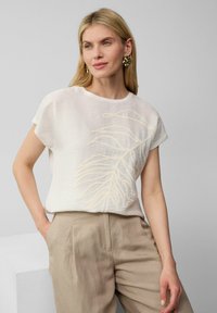 Haut blanc à manches courtes en tissu texturé avec un subtil motif de feuilles beige. Associé à un pantalon beige clair, ample.