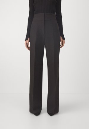 Femme portant un pantalon noir taille haute à jambes larges et un haut noir ajusté à manches longues, debout devant un fond clair uni.