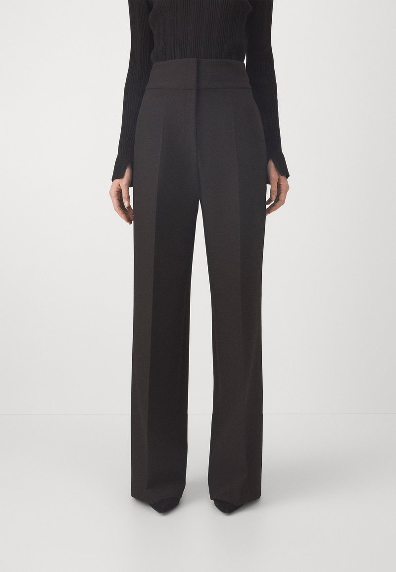 Femme portant un pantalon noir taille haute à jambes larges et un haut noir ajusté à manches longues, debout devant un fond clair uni.