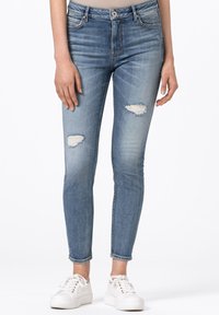 Kvinna i ljusblå rivna trånga jeans och vita snörskor, stående mot en enkel vit bakgrund.