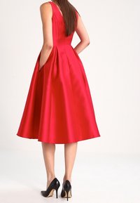 Robe A-line en satin rouge avec un design sans manches, jupe plissée et poches latérales, assortie à des escarpins noirs.