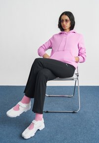 Lys rosa hettegenser med snorer, svarte bukser og hvite joggesko med rosa detaljer. Modell sitter på en kromstol mot en ensfarget bakgrunn.