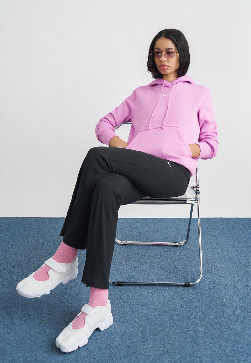 Lys rosa hettegenser med snorer, svarte bukser og hvite joggesko med rosa detaljer. Modell sitter på en kromstol mot en ensfarget bakgrunn.