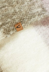 Uldblandings tæppe med et abstrakt mønster i nuancer af beige og creme, struktureret overflade og et lille logo-label i brunt læder.