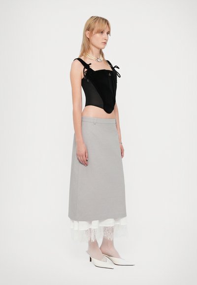 Νέα γυναίκα που φο�ράει μαύρο αμάνικο τοπ με φιόγκους, γκρι μεσοφόρι με λευκή δαντελένια φάσα, διάφανες λευκές καλσόν και λευκά μυτερά τακούνια.