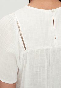 Hvid bluse med tekstureret stof, korte ærmer og en knaplukning bagpå. Har et subtilt rutenetmønster og blonde detaljer.