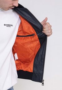 Zwarte leren jas met een oranje gewatteerde voering en een ritszak aan de binnenkant. Heeft een geribde zoom en een ritsluiting.