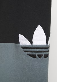 Svart och grå bomullst-shirt med en vit broderad logotyp med tre blad och svarta randdetaljer nära fållen.