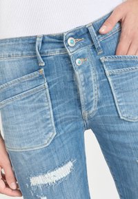 Jeans en denim bleu clair avec taille haute, double fermeture à bouton avec des accents turquoise et détail effiloché aux genoux. Quatre poches visibles.