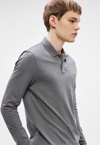 Graues, langärmeliges Polohemd mit Kragen, ausgestattet mit zwei Knöpfen und einem kleinen Logo auf der Brust, gefertigt aus glattem Stoff mit einer dezenten Textur.
