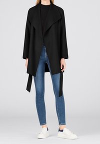 Cappotto nero avvolgente con ampio colletto, apertura frontale e legature laterali, abbinato a jeans blu aderenti e sneakers bianche. Texture liscia e design minimale.