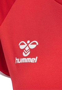 Punainen urheilupaita, jossa on sileä pinta ja valkoinen "hummel"-logo sekä tyylitelty emblem etupuolella. Klassinen urheilullinen muotoilu.