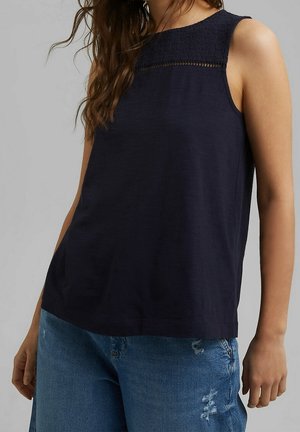 Frau trägt ein ärmelloses marineblaues Top mit Spitzenbesatz am Ausschnitt, kombiniert mit zerfetzten blauen Jeans.