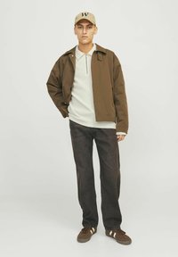 Chaqueta ligera marrón sobre una camisa de rayas blanca, combinada con vaqueros oscuros texturizados y zapatillas marrones con franjas blancas. Gorro beige con logo.
