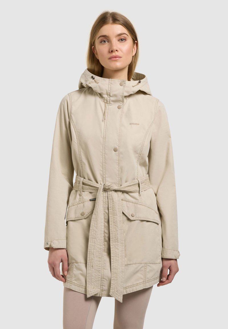 Femme portant une veste à capuche beige avec ceinture et grandes poches avant, debout devant un fond gris clair.