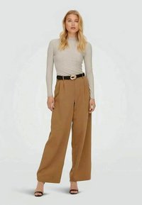 Camisola beige de mangas longas, calças largas castanhas de cintura alta, cinto preto com fivela dourada, usando sandálias de salto pretas, fundo minimalista.