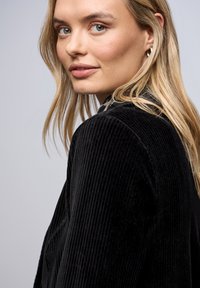 Veste en velours côtelé noir avec des côtes verticales, dotée d'un col. Le mannequin a de longs cheveux blonds. L'arrière-plan est constitué d'un dégradé doux.