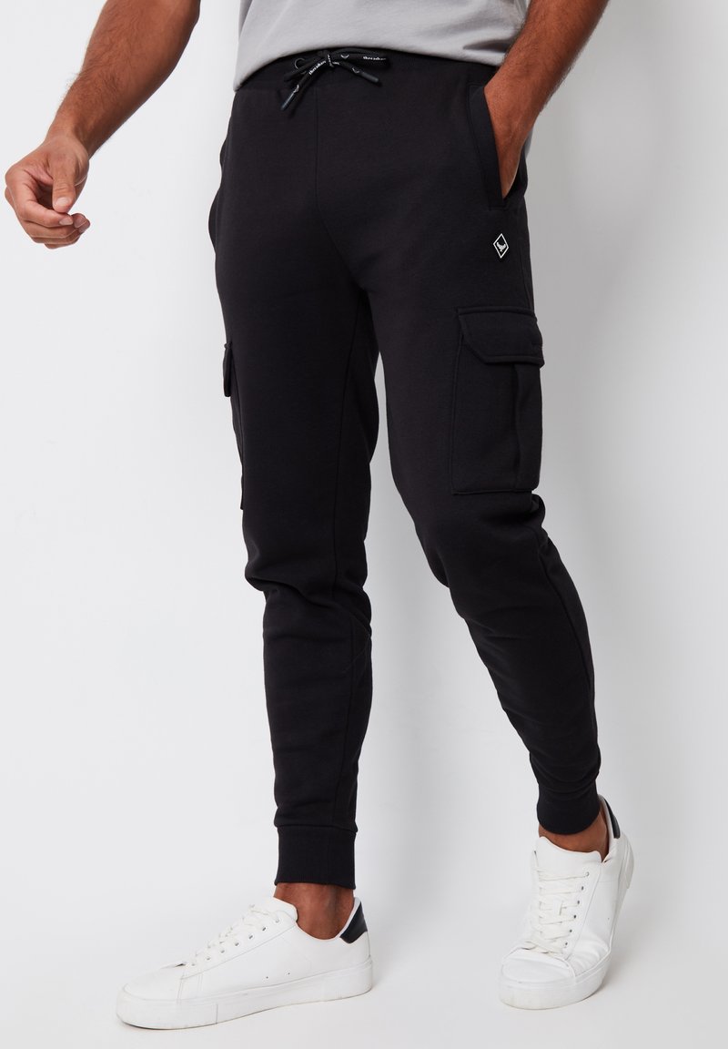 Uomo che indossa pantaloni jogger cargo neri con tasche laterali e sneakers bianche, in piedi davanti a uno sfondo bianco.