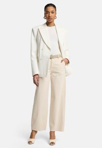 Weißes doppelt geknöpftes Blazer, gerippter weißer Oberteil, hellbeige weit geschnittene Hose, verzieller Gürtel und offene Pumps. Einfaches, klares Design.