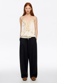 Kremowa jedwabna koszulka na ramiączkach z koronkową lamówką i wiązaniem z przodu, zestawiona z luźnymi czarnymi spodniami i brązowymi wsuwanymi butami.