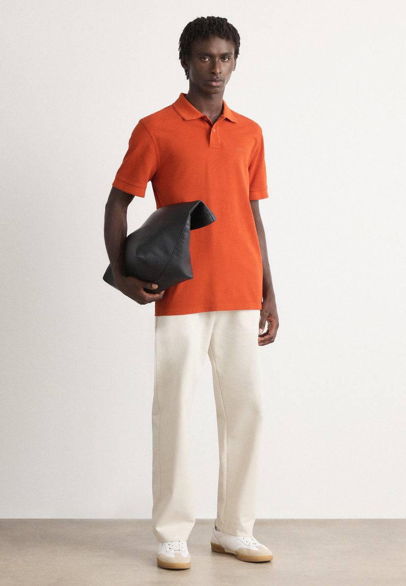 Homme portant un polo orange et un pantalon blanc, tenant un grand sac en cuir noir plié, se tenant sur un sol gris contre un mur blanc.