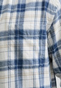 Blau-weiß kariertes Hemd aus Stoff, mit einer Tasche, und einem Rautenmuster in verschiedenen Blautönen und hellbraunen Akzenten.