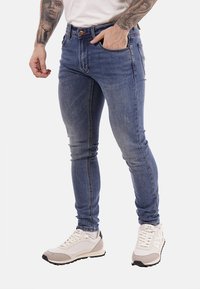 Uomo che indossa jeans blu aderenti, sneakers bianche e una t-shirt bianca, con braccia tatuate e una mano in tasca.