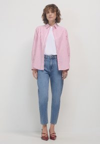 Frau trägt ein rot-weiß gestreiftes Hemd über einem weißen T-Shirt, blaue hochtaillierte Jeans und rote spitze Absätze und steht vor einem weißen Hintergrund.