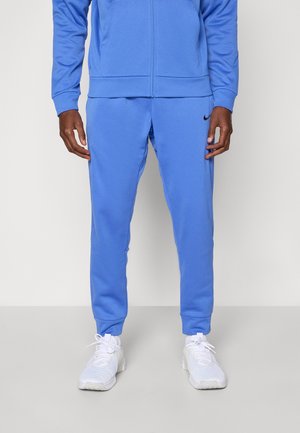 Vue recadrée d’un mannequin portant un survêtement bleu composé d’une veste zippée, d’un pantalon de jogging et de baskets blanches.