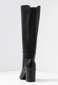 Bota negra hasta la rodilla con un exterior de cuero suave, paneles laterales elásticos y un tacón robusto y acanalado. Diseño sencillo con punta afilada.