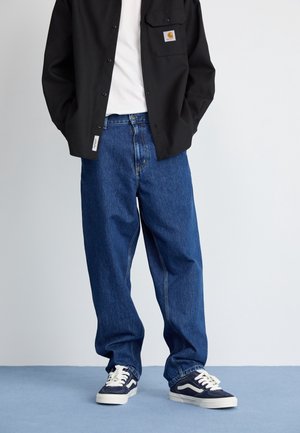 Person wearing loose blue jeans, black button-up shirt with logo patch on pocket, white t-shirt, and black-and-white sneakers standing on blue floor.